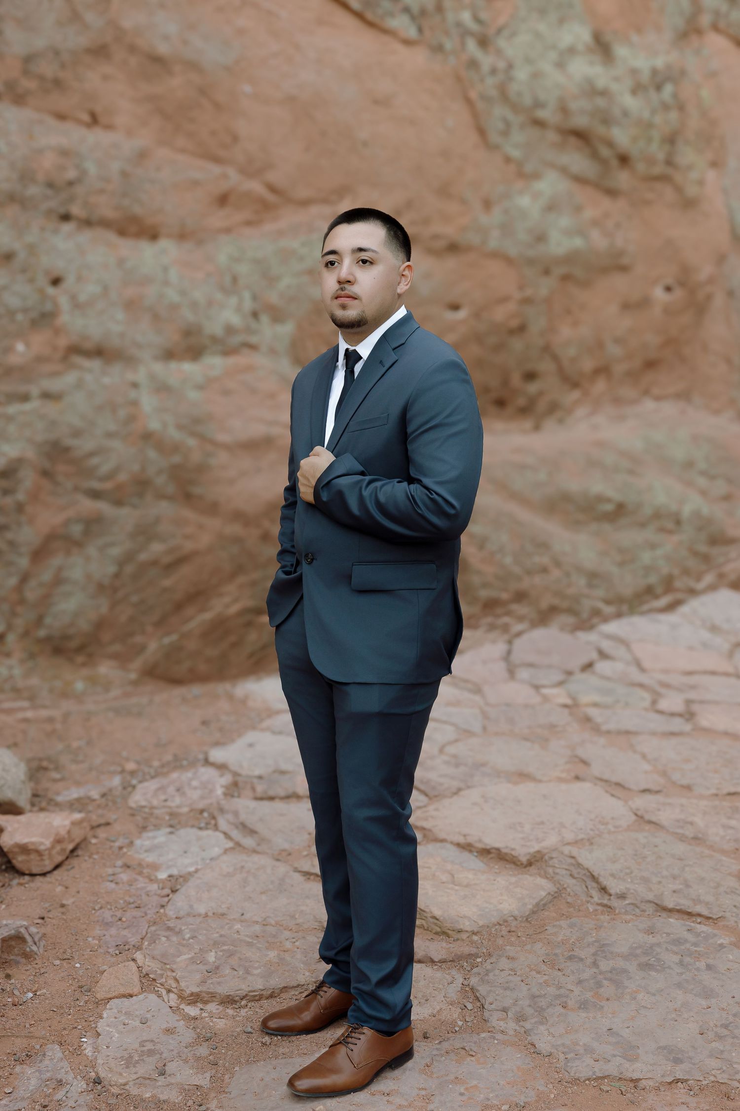 Groom in dark suit and brown shoes standing on rocky terrain against red rock formations.
