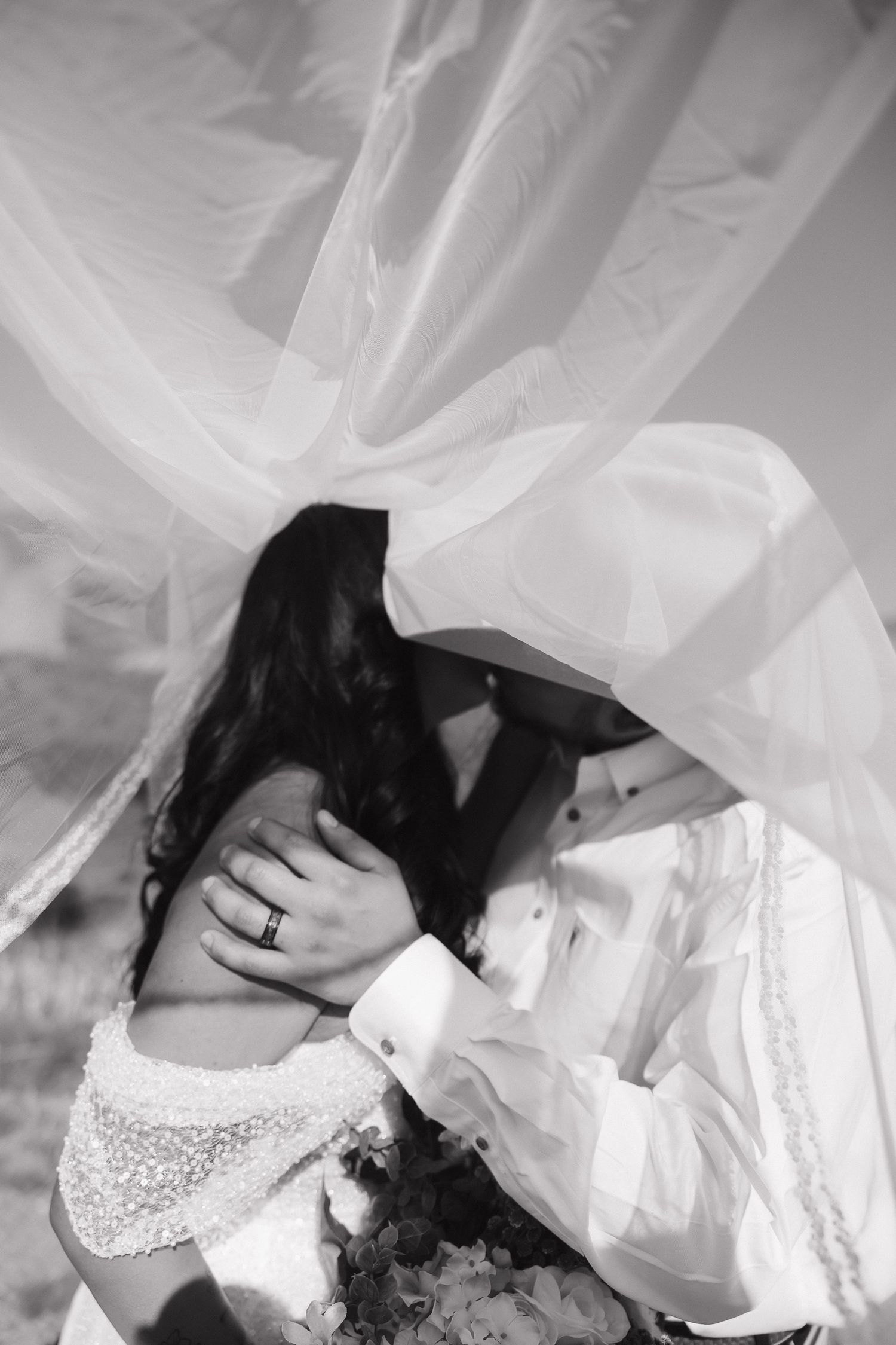 A romantic black and white wedding photo showing a couple embracing beneath flowing white veils and fabric.