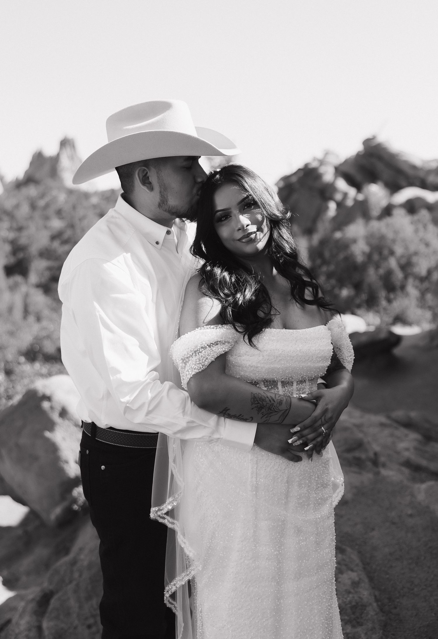 A romantic black and white wedding photo featuring a couple embracing outdoors in western attire.
