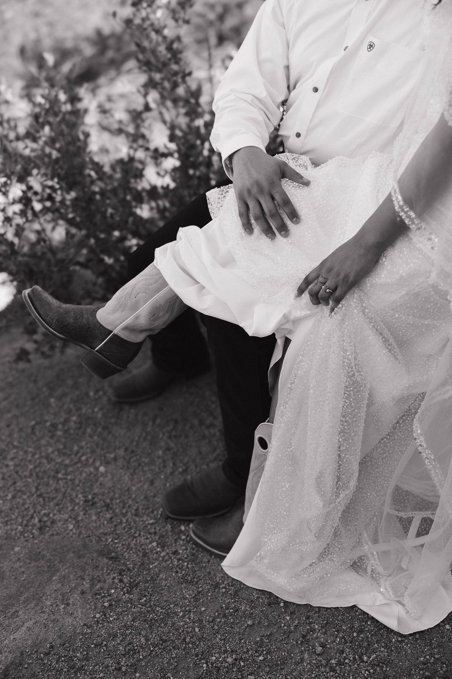Black and white detail shot of cowboy boots and a flowing wedding dress hem against natural terrain.