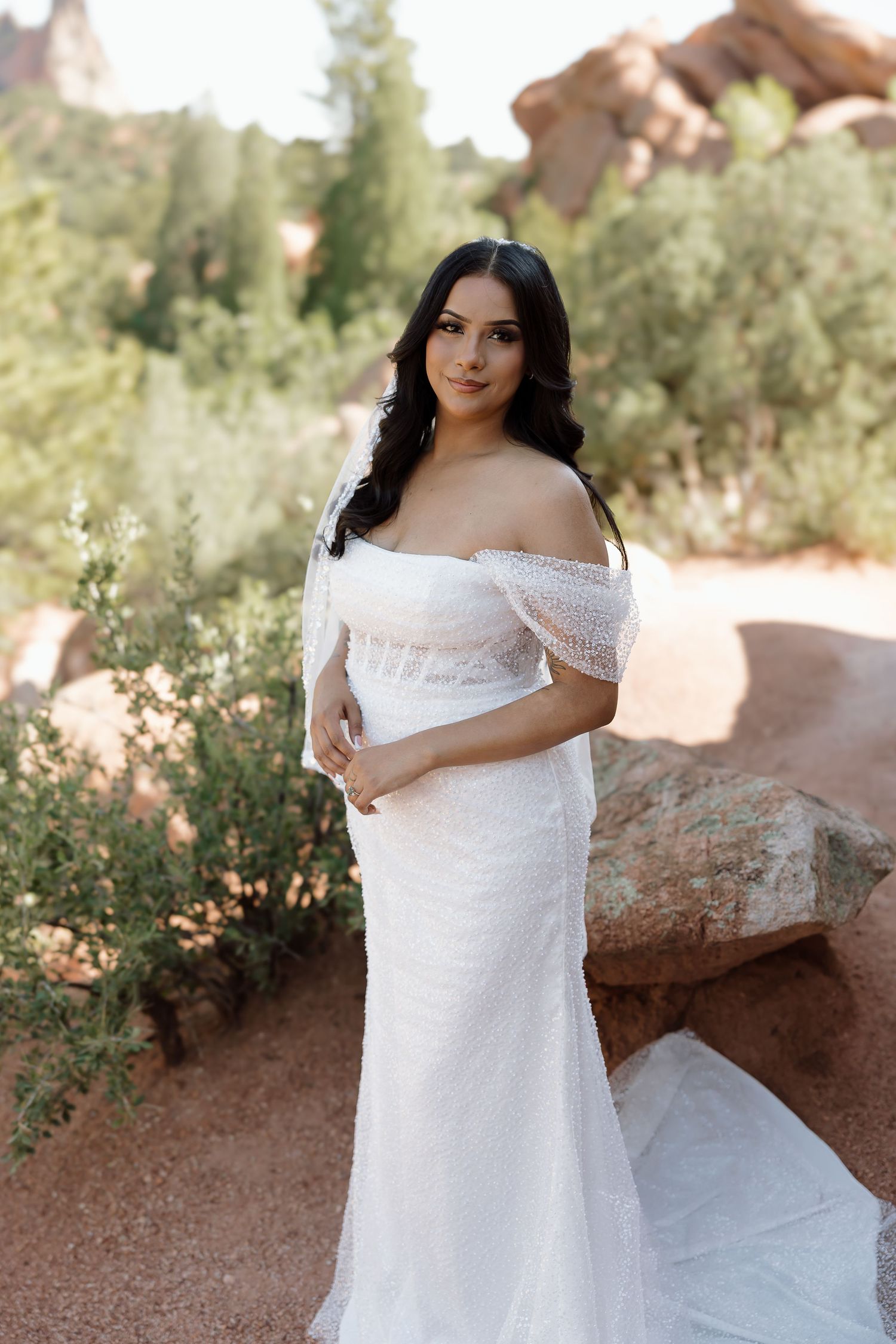 A bride in a white off-shoulder wedding dress poses elegantly against rustic red rocks and desert foliage.