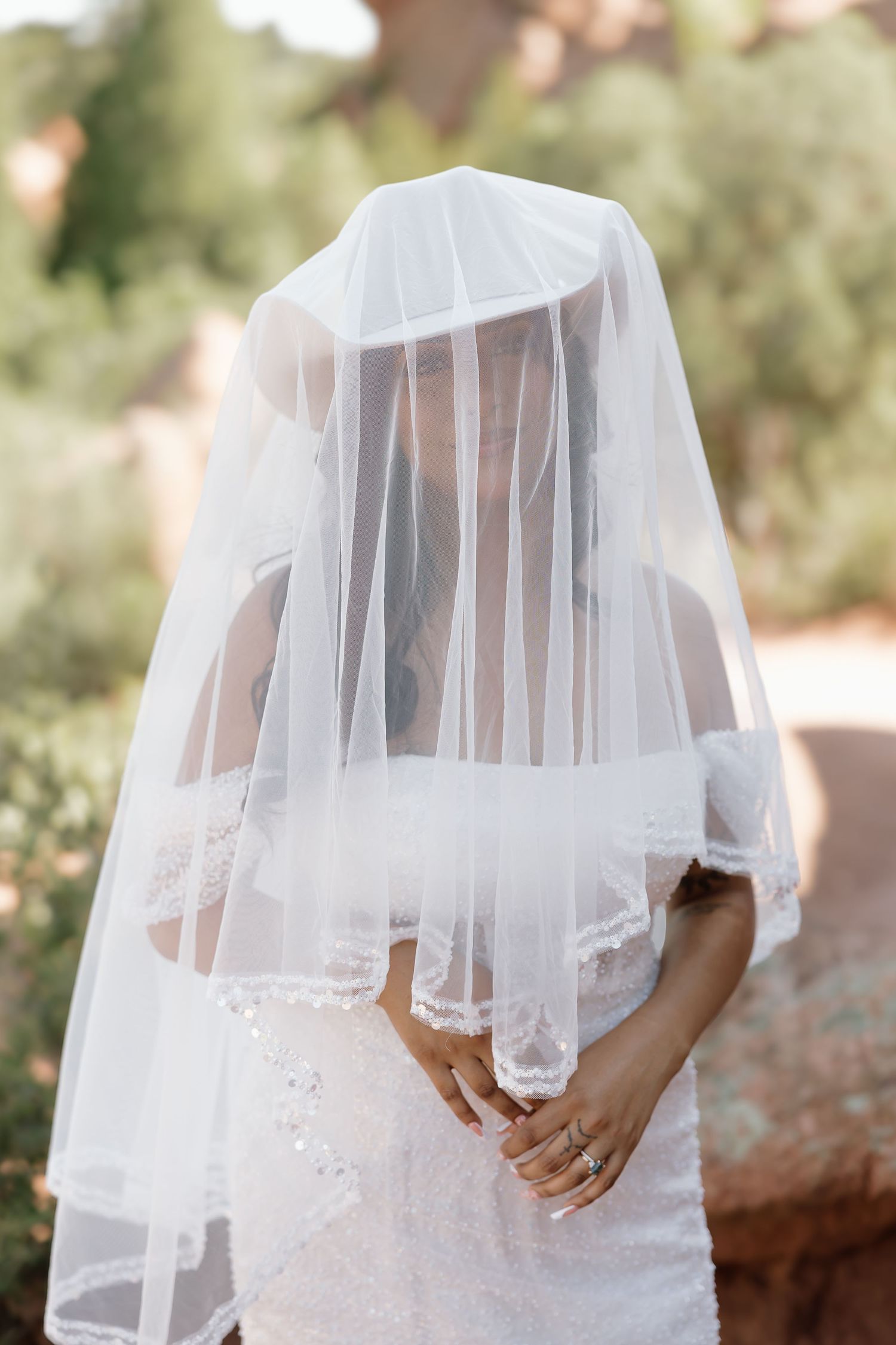 A bride wearing a white lace dress and delicate tulle veil stands in a sunlit garden setting.