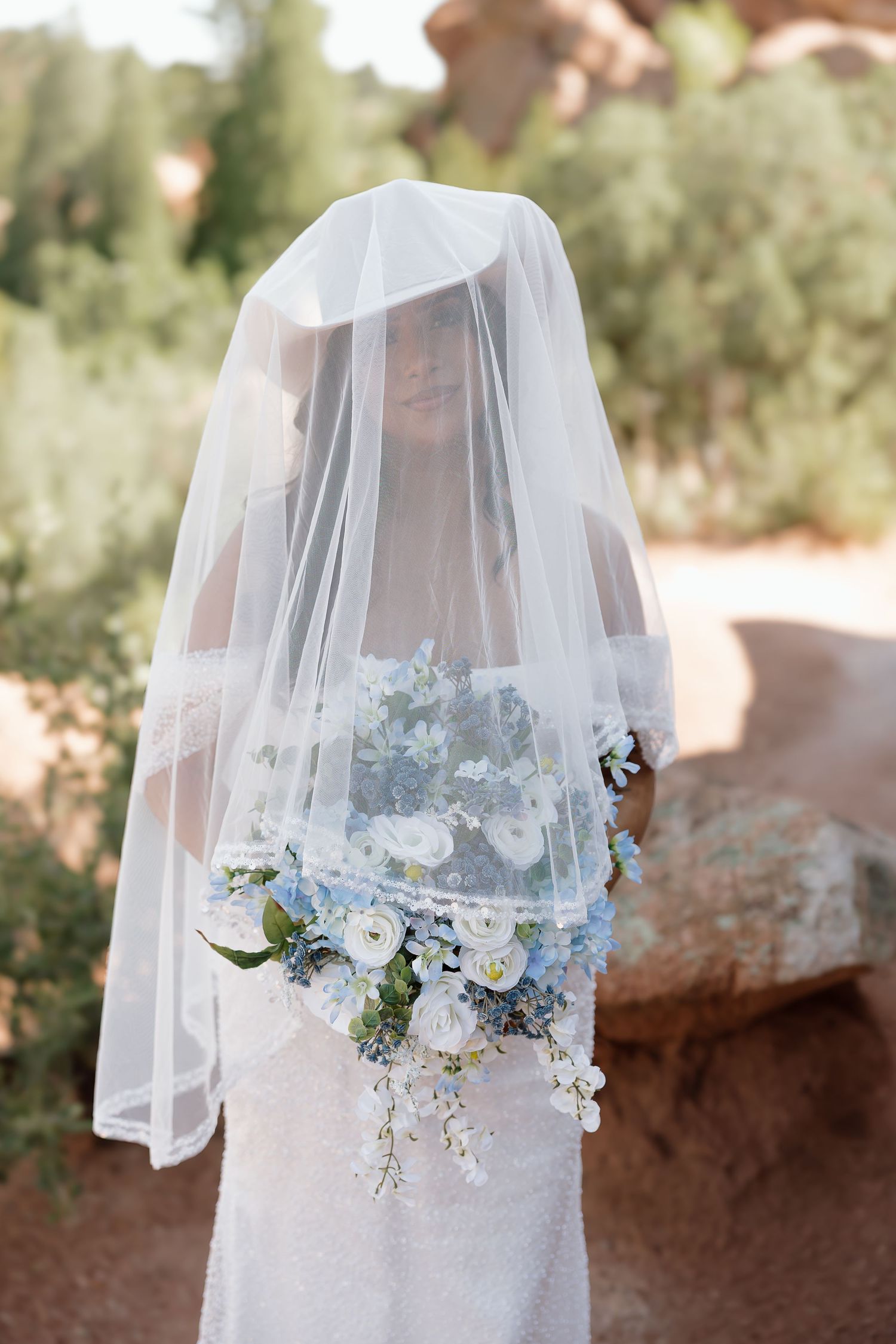 Ethereal bridal portrait featuring a delicate white veil and cascading blue and white floral bouquet in a garden setting.