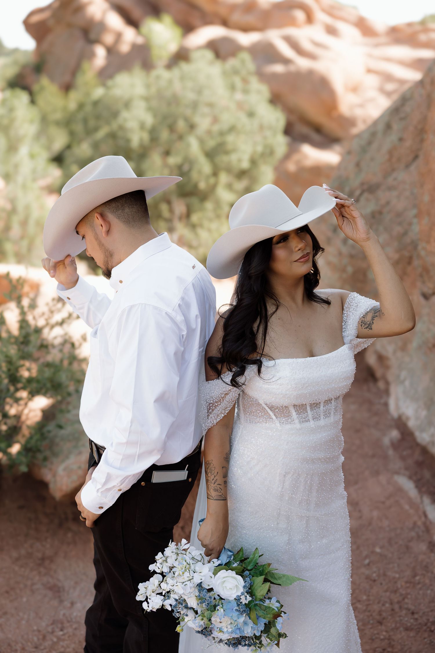 A western-themed wedding couple in white cowboy hats pose against red rocks with a blue and white bouquet.