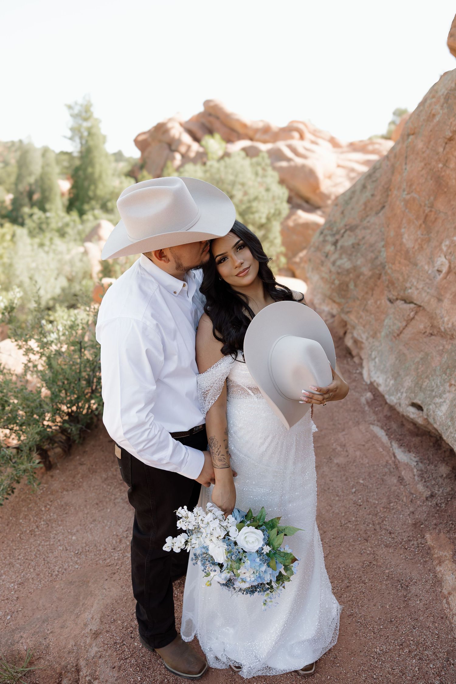 A couple in a western-style wedding shoot poses intimately against red rock formations in a desert landscape.