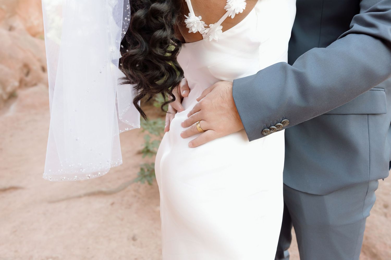 Close-up photo showing hands gently embracing figure in white wedding dress with soft curled dark hair.