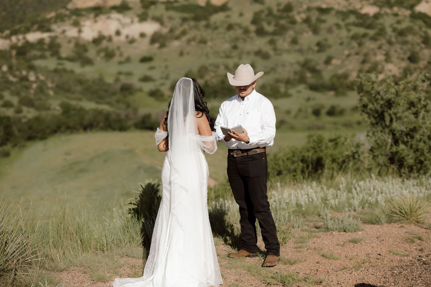 A couple in western wedding attire stands on a hilltop surrounded by green rolling meadows during their outdoor ceremony.