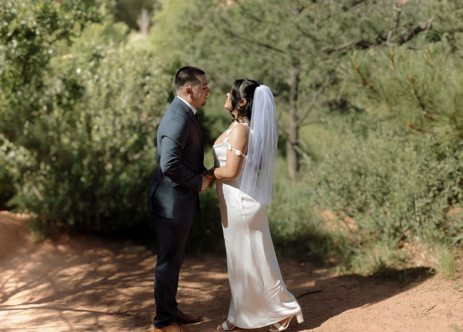 A couple shares a romantic moment in their wedding attire on a desert path with green foliage in the background.