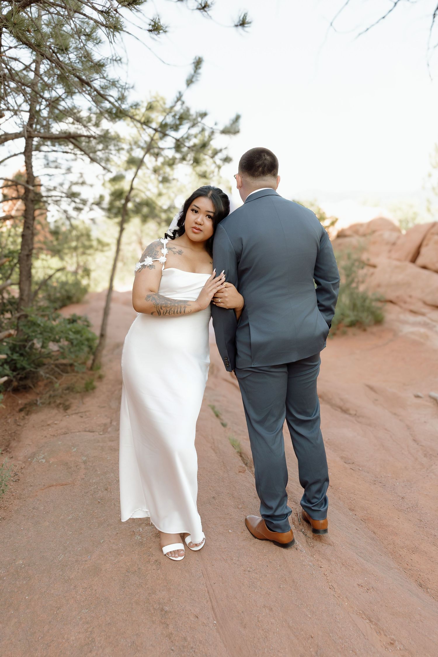A couple in formal attire walks together along a winding red rock path in nature during a serene photo session.