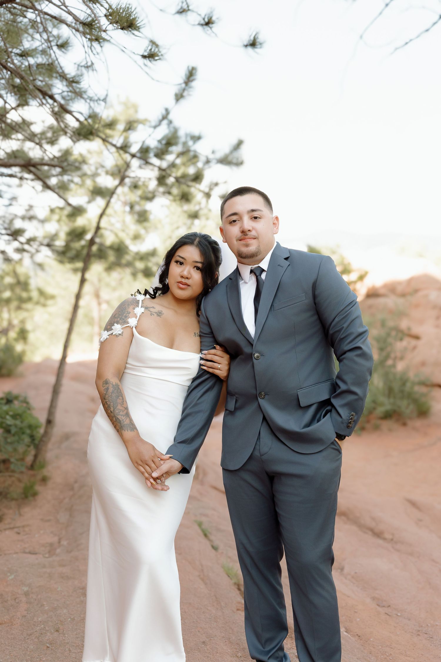 A romantic wedding photo of a couple in formal attire standing together on a desert path with pine trees in the background.