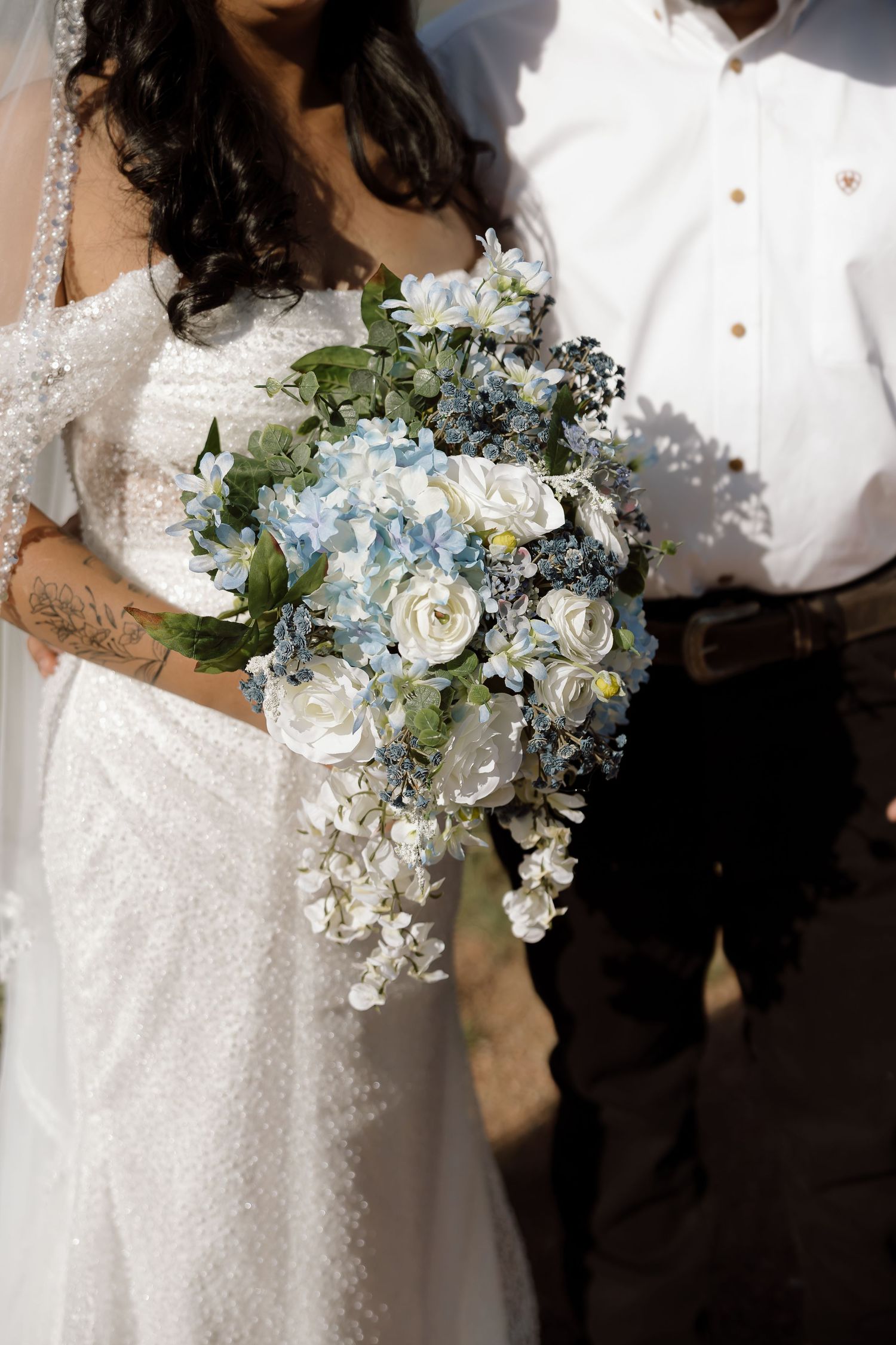 A cascading wedding bouquet features light blue and white flowers with greenery against a white lace wedding dress.