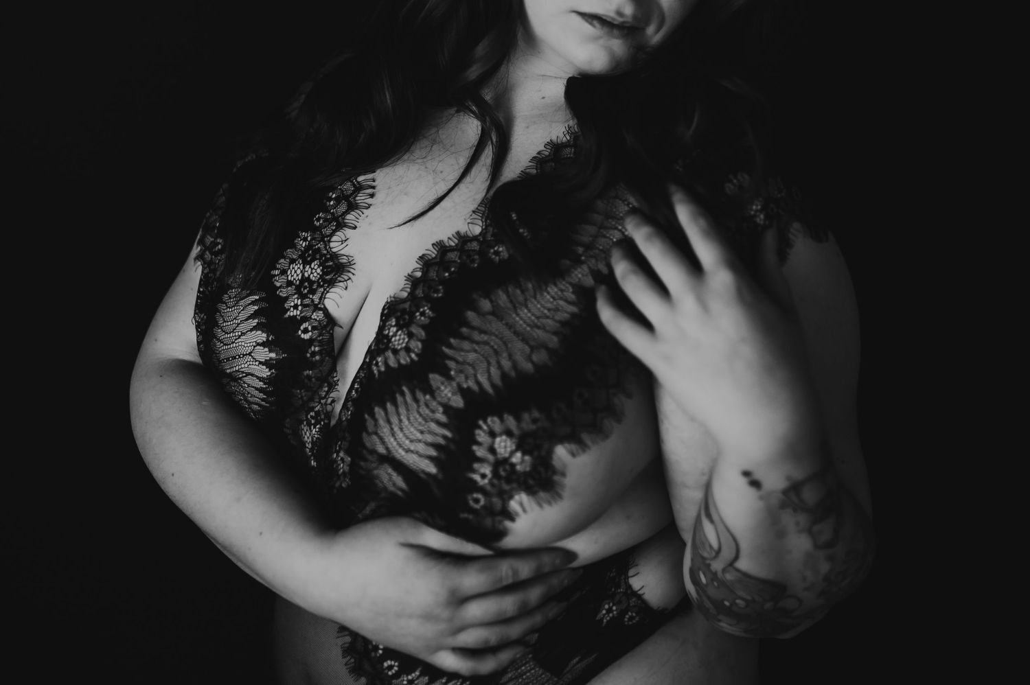 Black and white artistic portrait showing torso in lace lingerie with visible arm tattoo in dark moody lighting.