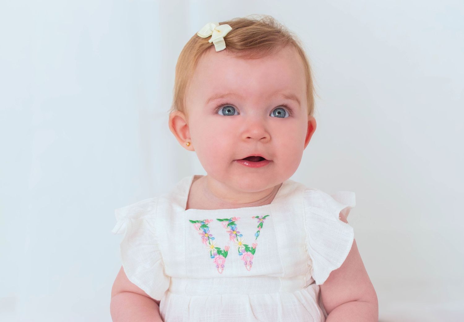 Baby in white dress with bow accessory against light background.