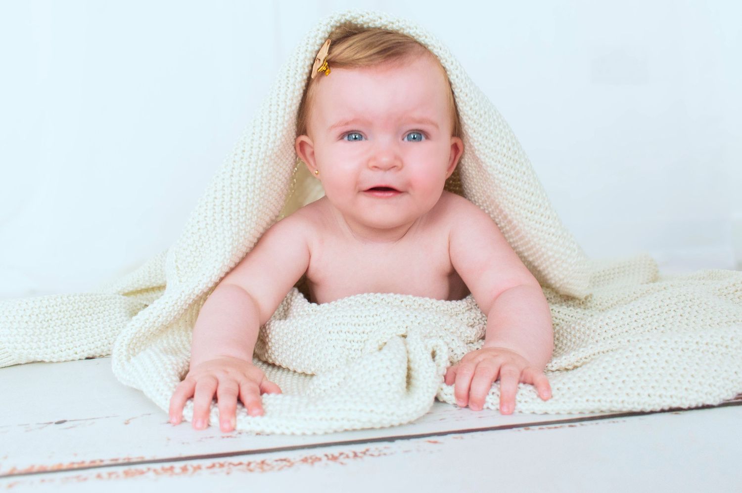 Baby peeks out from under white knit blanket during playful photo shoot.