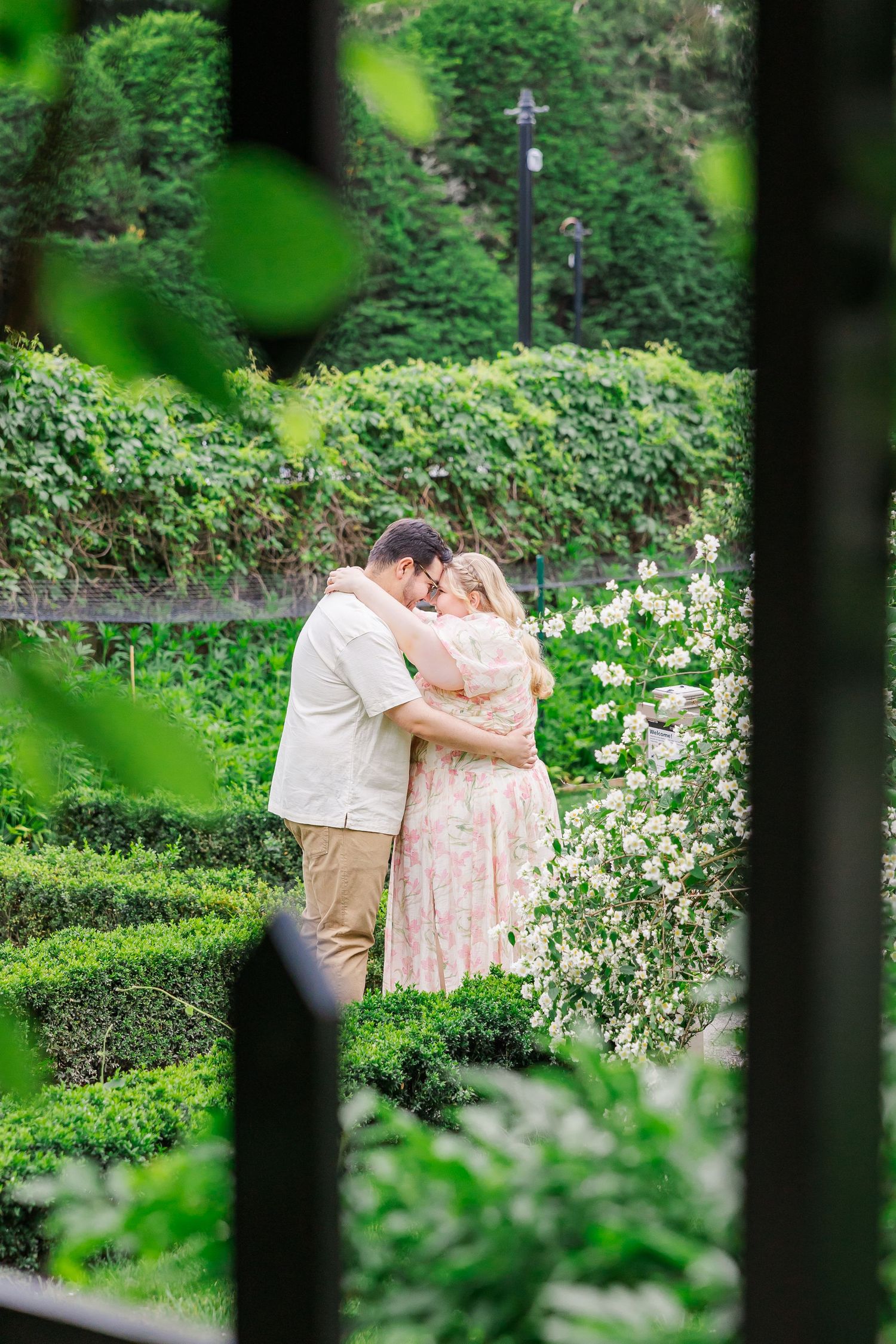 A romantic couple embraces in a lush green garden with blooming flowers and manicured hedges.