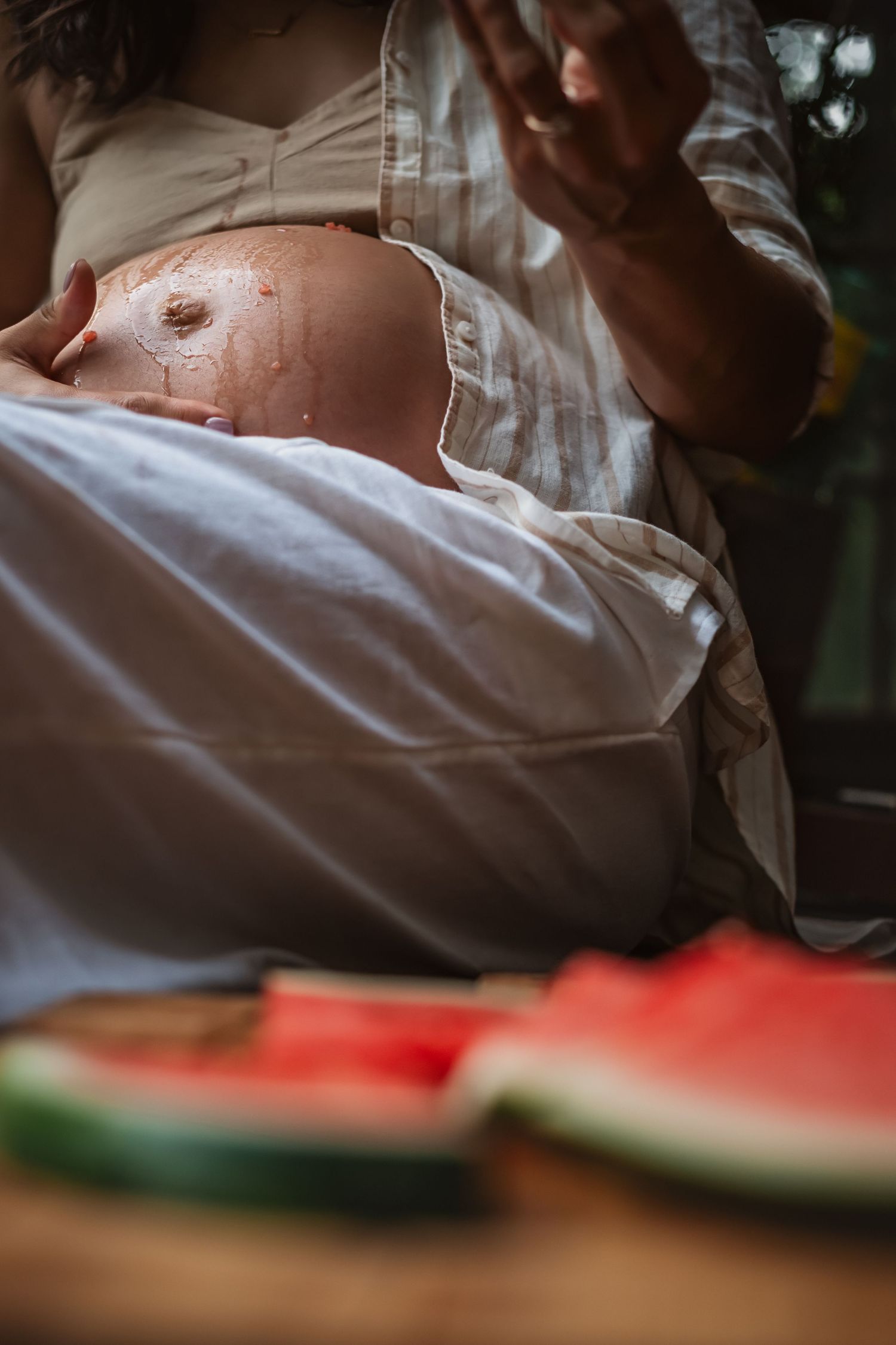 A pregnant woman in an open white shirt drips watermelon juice on her bare belly while she relaxes with sliced watermelon pieces in summer.