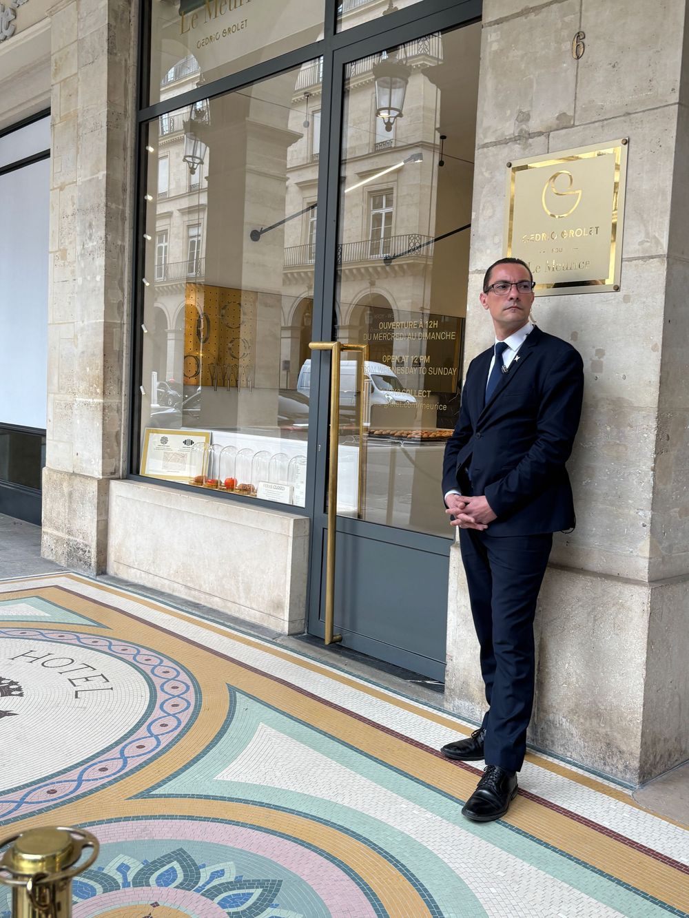 A person in a dark business suit stands in front of a luxury retail storefront with ornate mosaic floor tiles. Cedric Grolet pastry shop in.