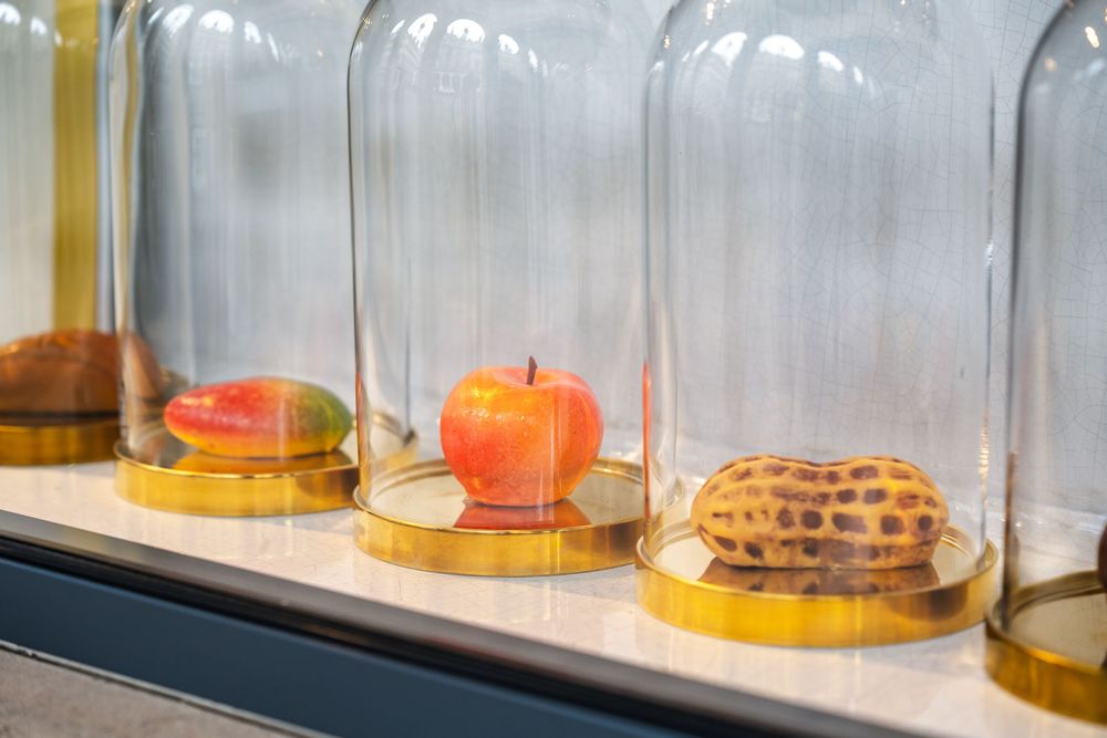 Multiple apples sit under clear glass cloches arranged in a row on a wooden shelf. Cedric Grolet Pasrties.