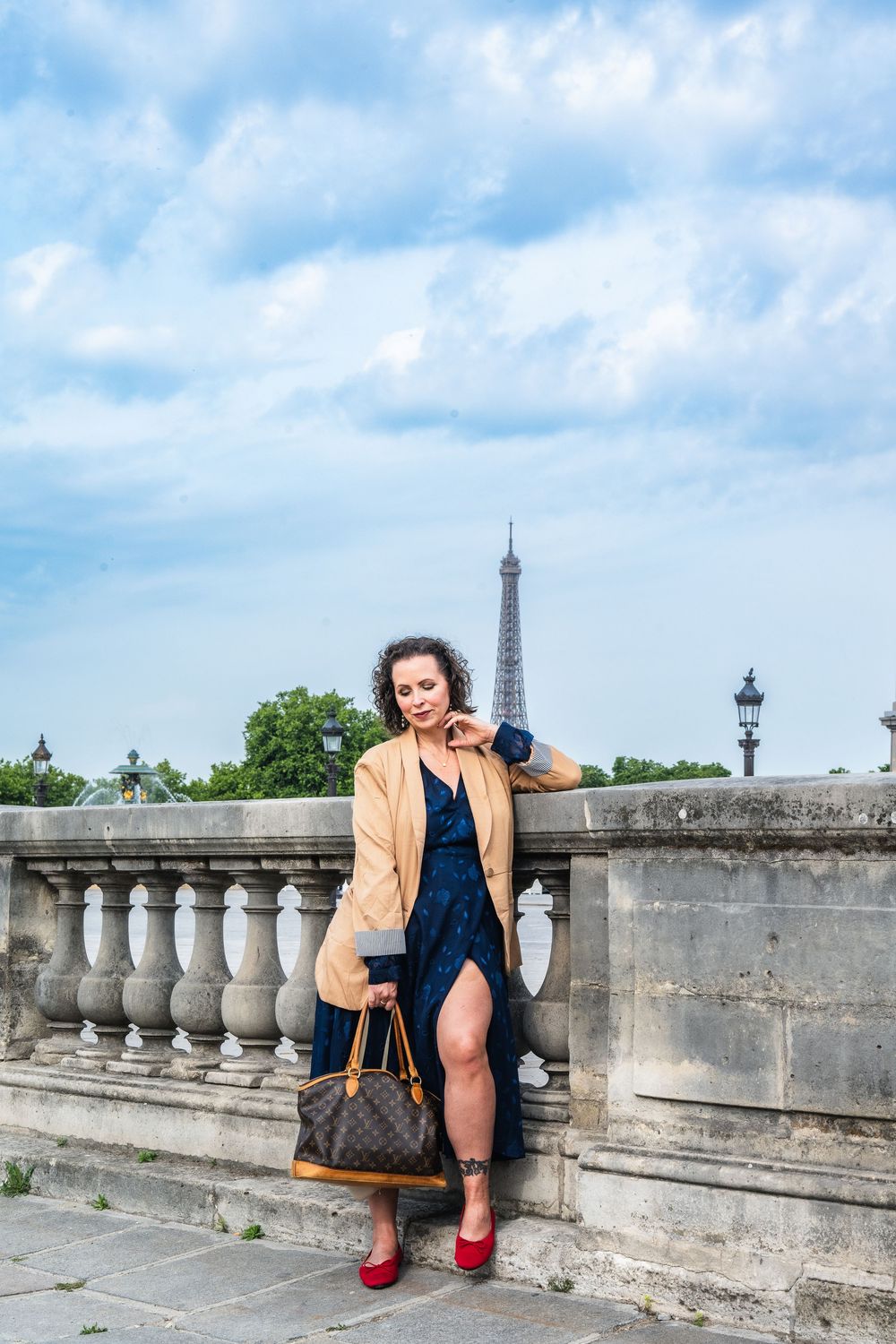 A person wearing a beige coat and navy dress poses on a stone balustrade with the Eiffel Tower visible in the background.
