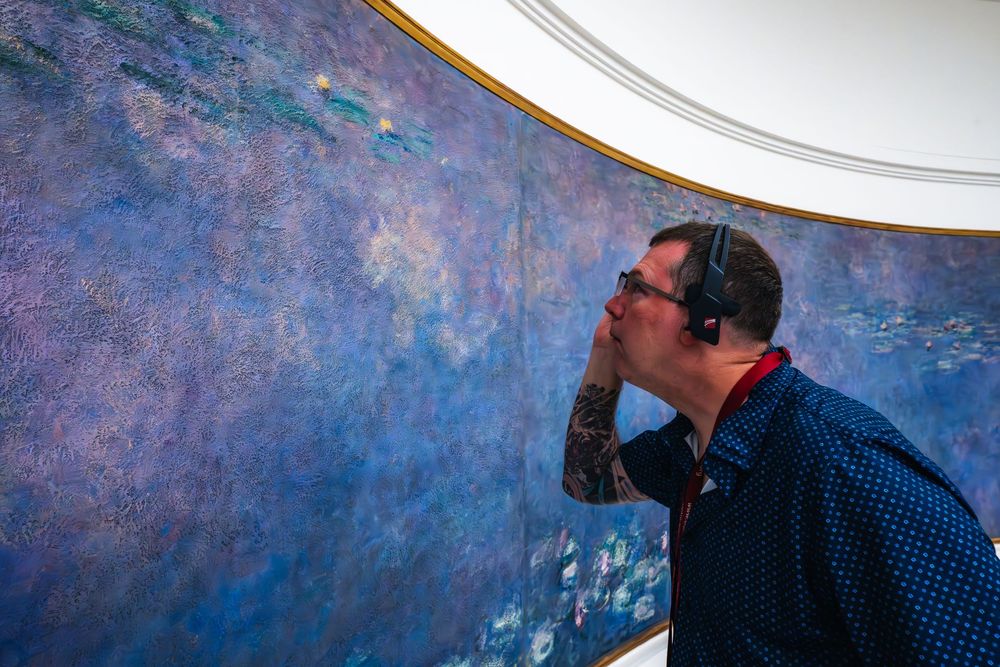 A person in glasses and polka dot shirt leans in to closely examine a large blue Monet water lily mural at a museum.