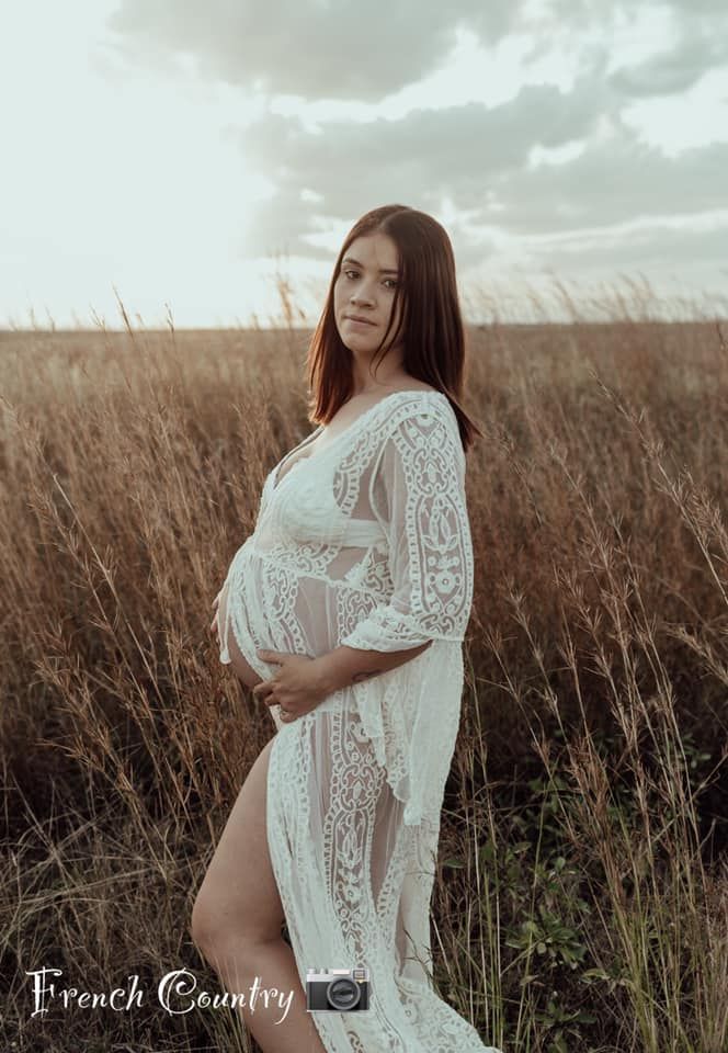 A figure in a flowing white lace dress poses gracefully in a tall grass field under moody cloudy skies.