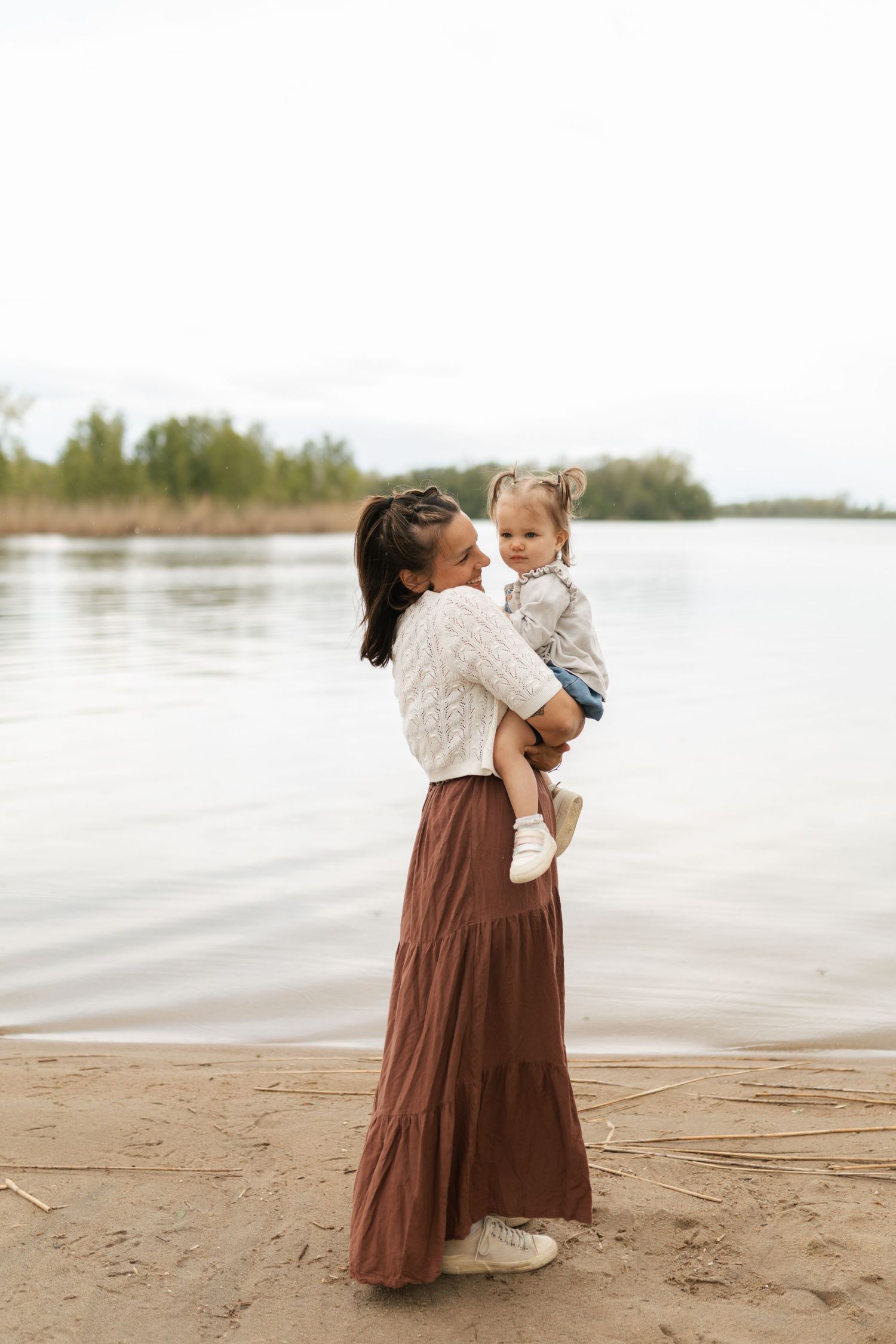 Mother holding toddler daughter by calm lake shore on overcast day.