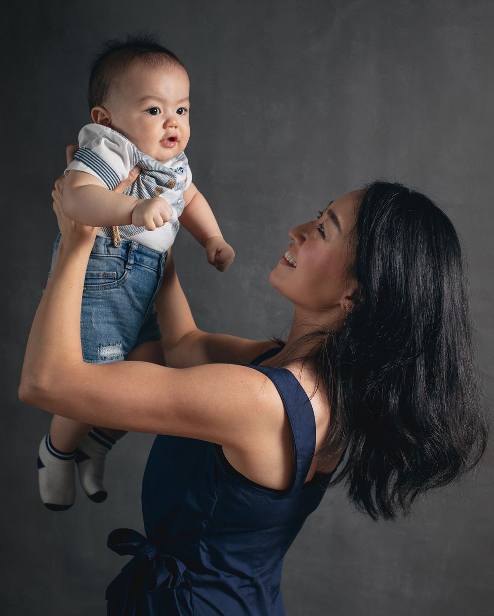 Mother in navy dress lifts and plays with a baby wearing overalls against a dark background.