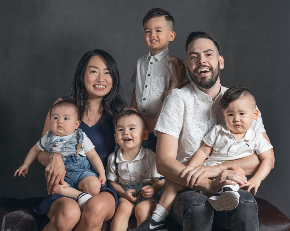A family with four young children poses for a studio portrait against a dark grey background.