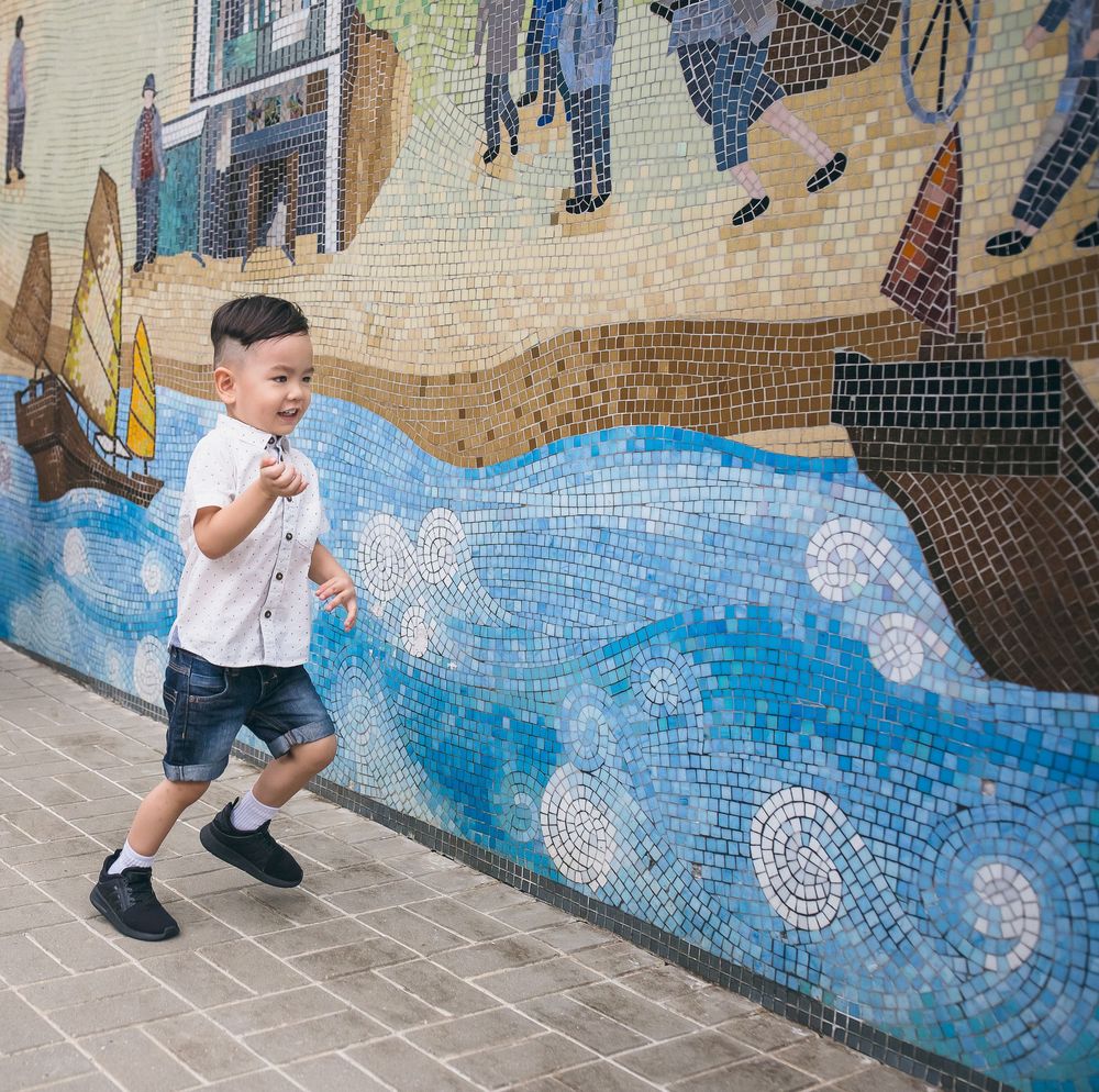 A child in a white t-shirt plays and poses in front of a blue mosaic wall mural on a stone walkway.