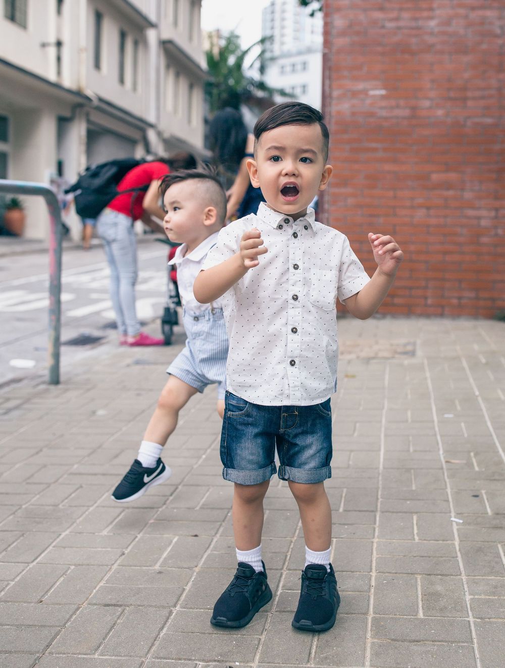 A toddler in a white polka dot shirt and denim shorts walks on a stone-paved outdoor pathway.