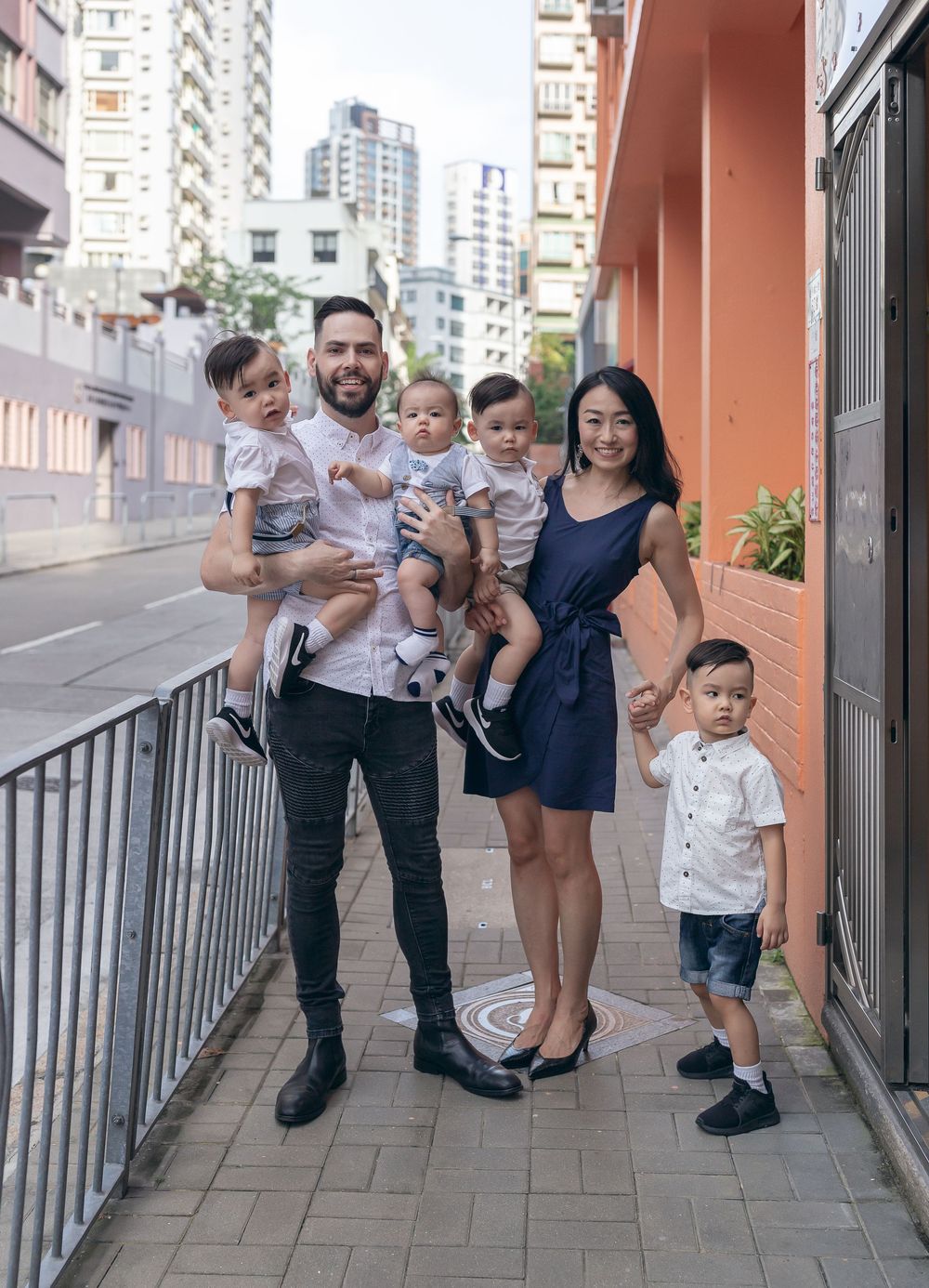A family poses playfully together on an outdoor walkway with orange walls in an urban setting.