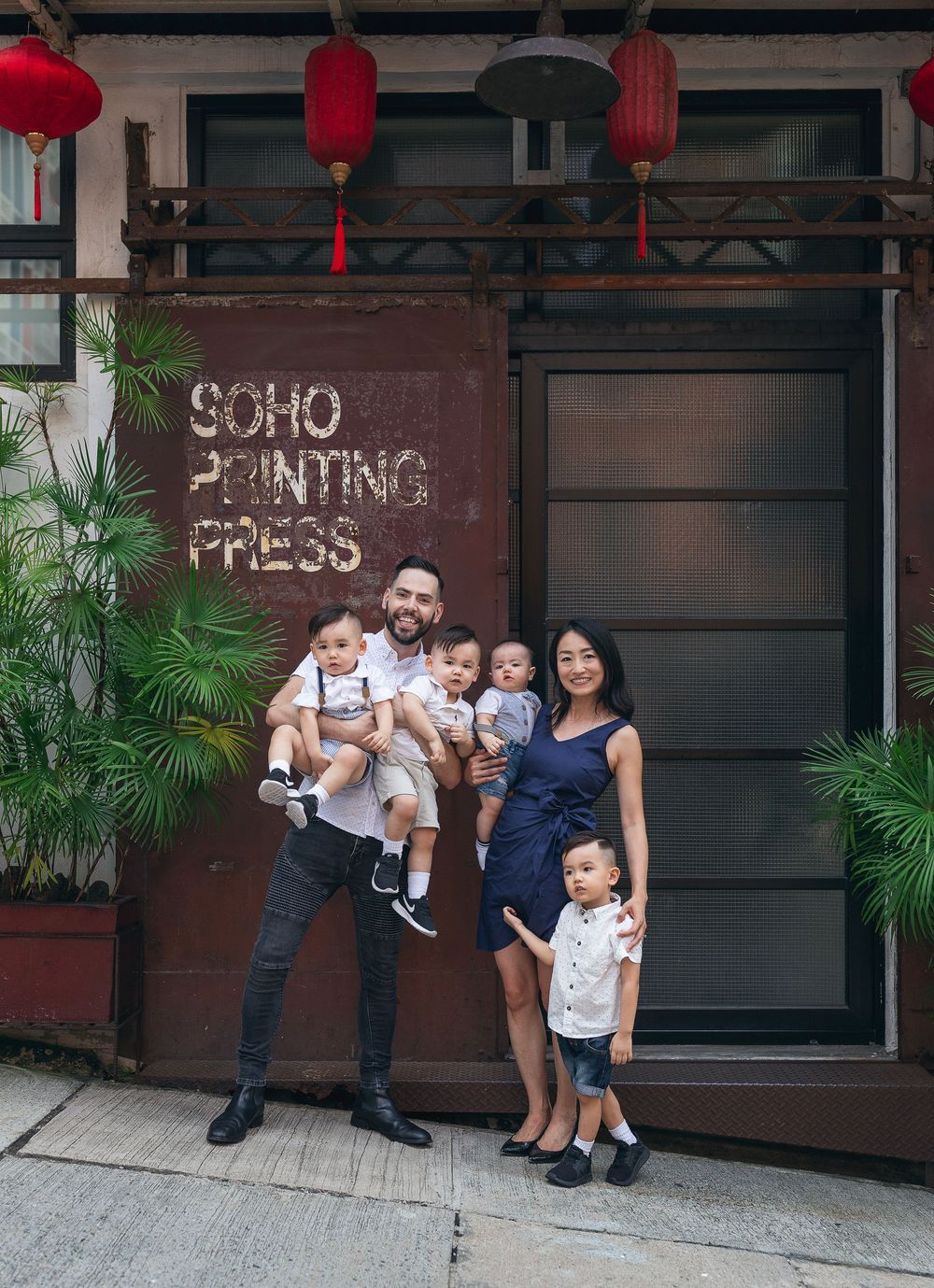 A family poses outside a building with red lanterns and a Soho Printing Press sign surrounded by plants.
