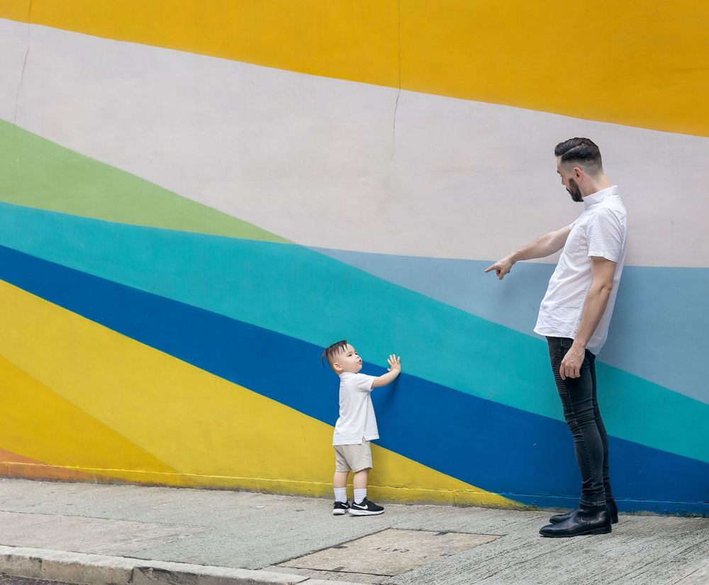Adult and small child interact playfully against a colorful striped wall with yellow, teal and blue geometric shapes.