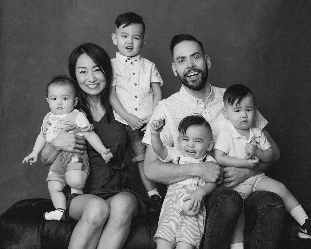 Black and white studio portrait of a family with four children sitting together and smiling against a neutral backdrop.