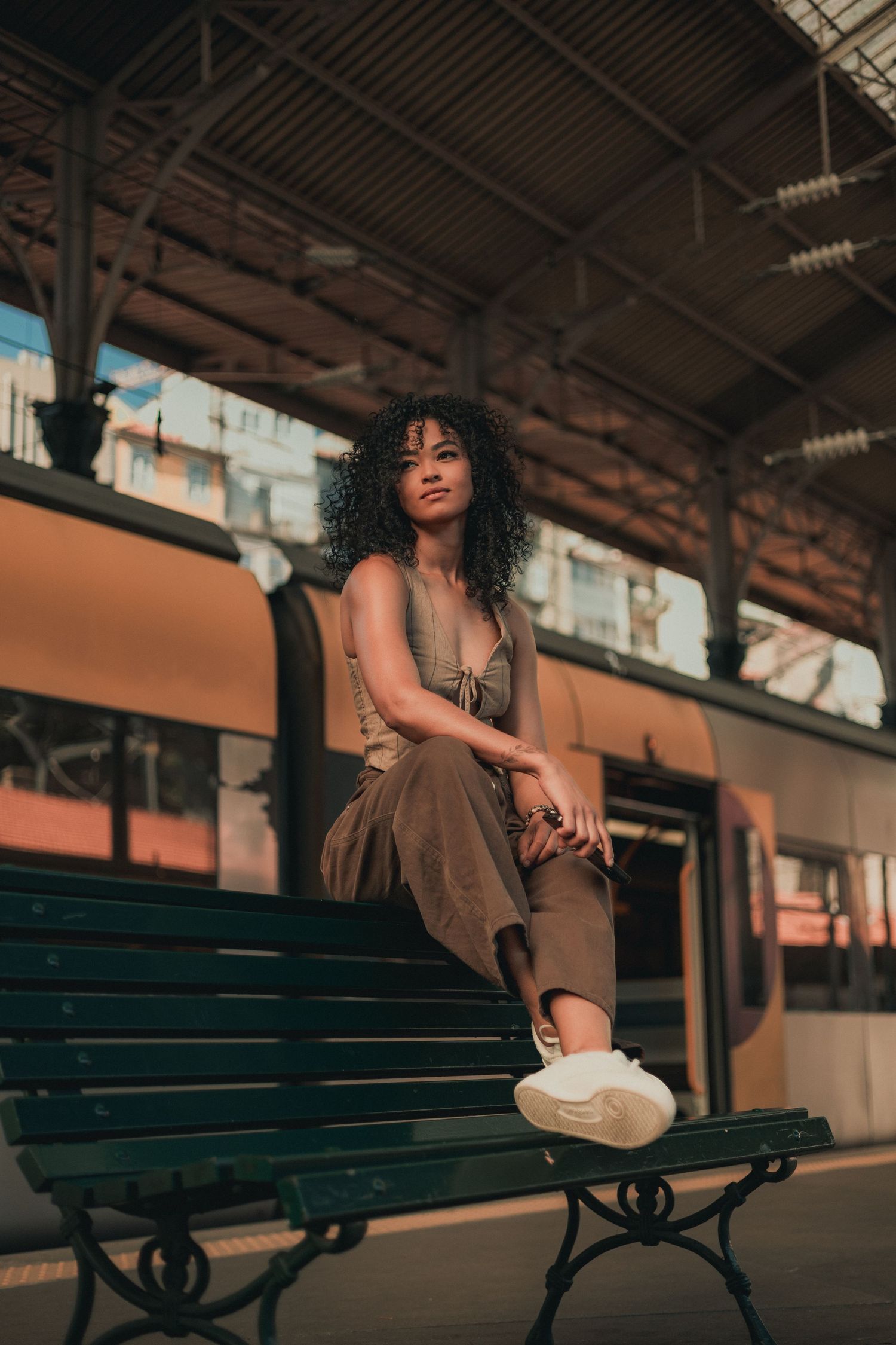 Curly-haired model in brown outfit poses elegantly on vintage train station bench with a hat at sunset.