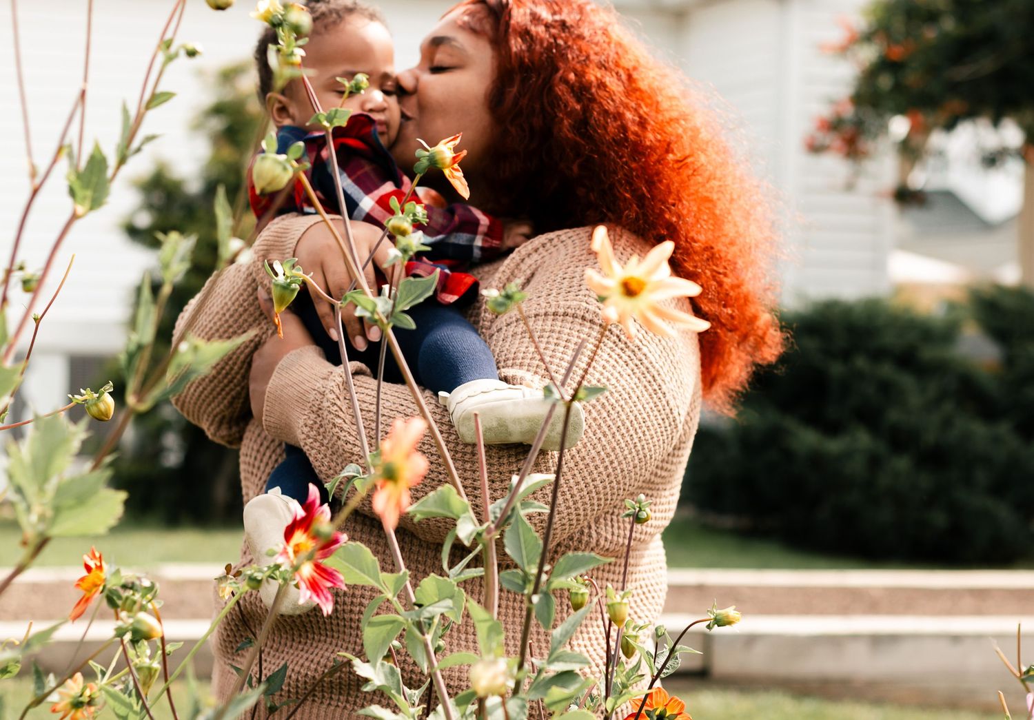 Person with curly red hair wearing a beige sweater smelling flowers in a sunny garden.