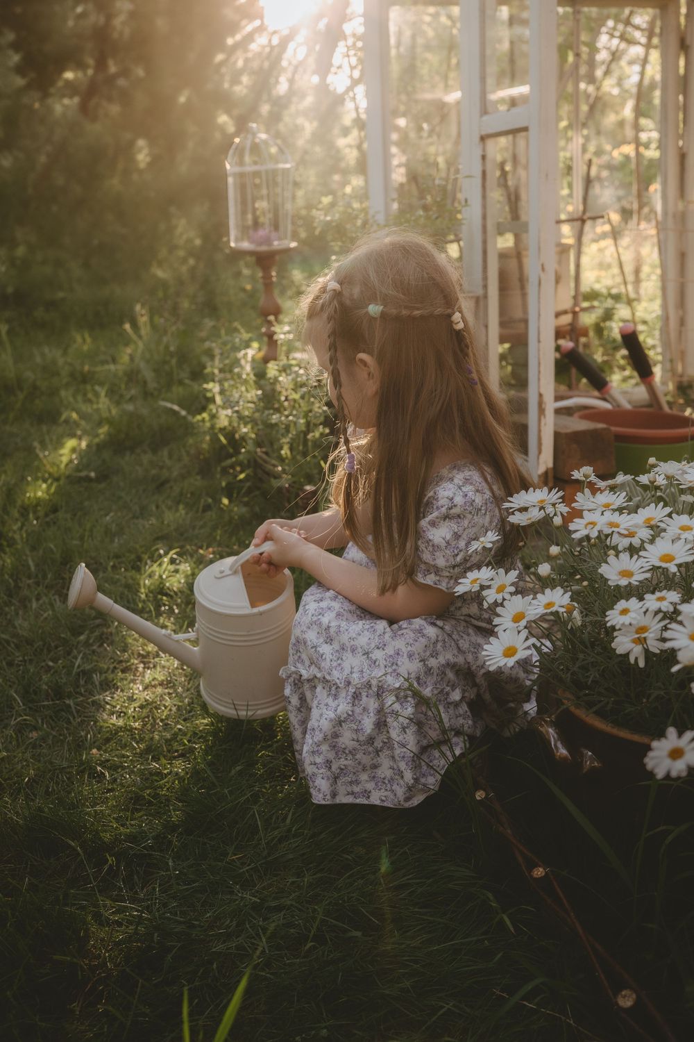 A young child in a floral dress tends to garden flowers with a watering can in golden sunlight.
