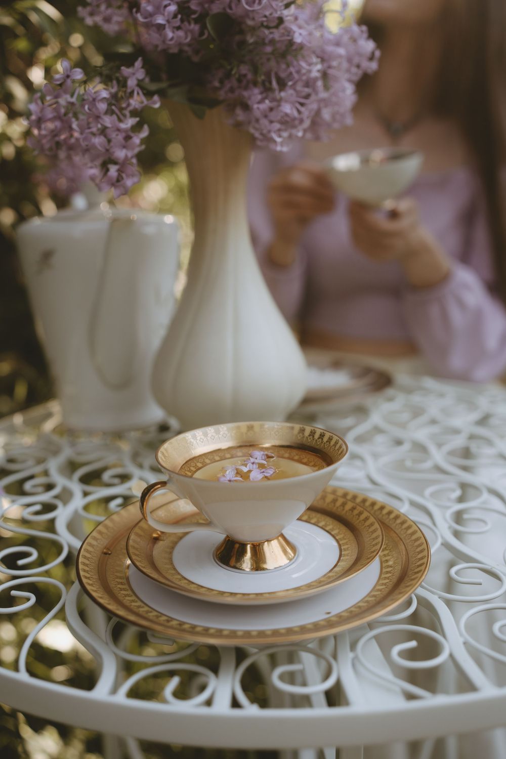 Vintage teacup with gold trim on white lace tablecloth next to lilac bouquet in white vase.