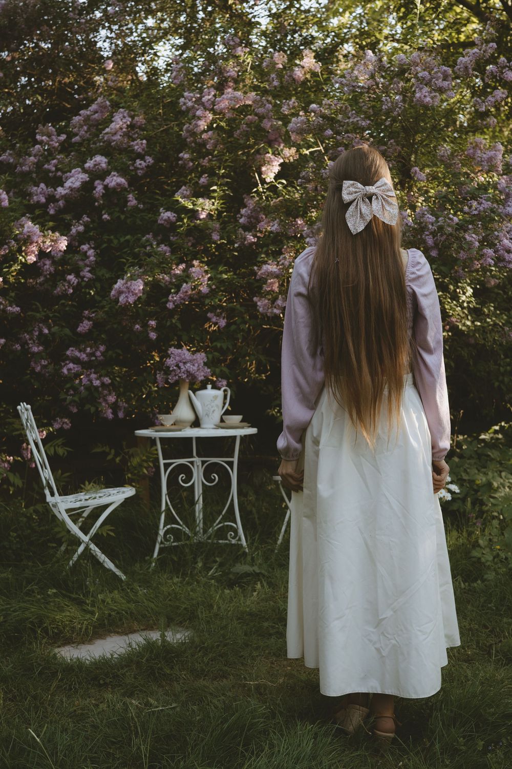 Vintage garden scene with white tea table beside blooming lilac bushes and a long-haired figure in white dress.