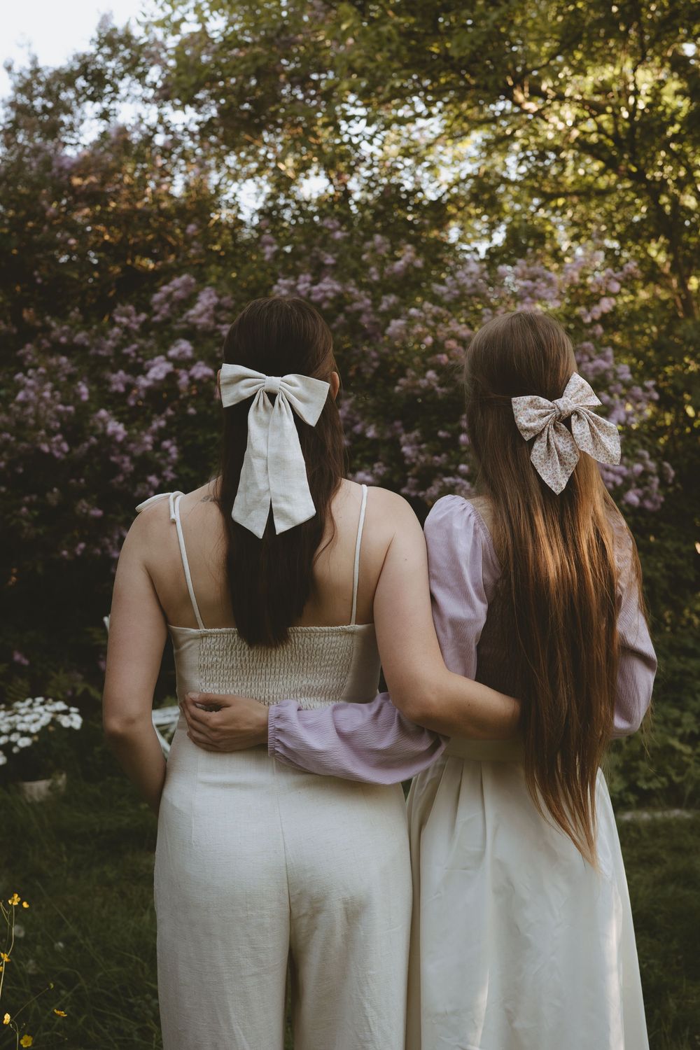 Two women in white dresses with matching white bows in their hair stand together in a garden at sunset.