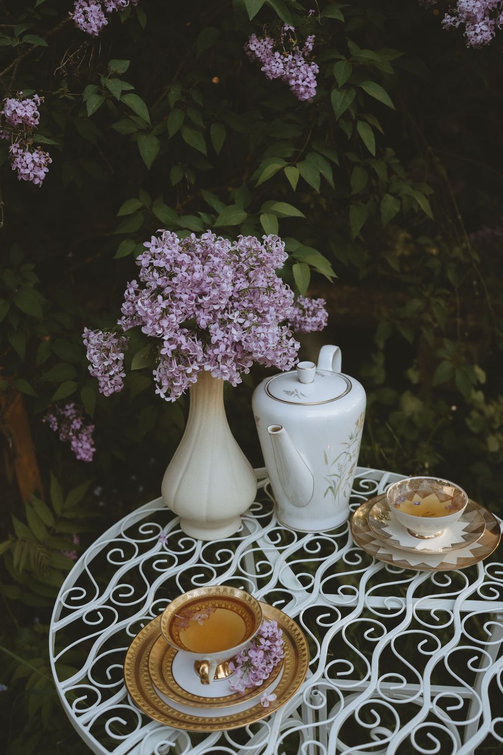 Elegant tea service with lilacs on white wrought iron table features gold-rimmed teacups.