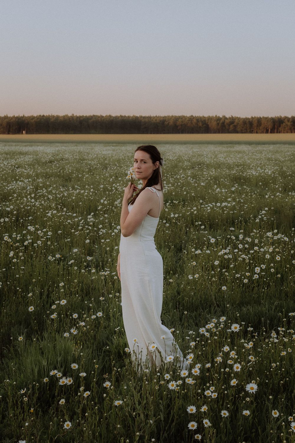 A person in a white dress stands thoughtfully in a blooming meadow at sunset with wildflowers all around.