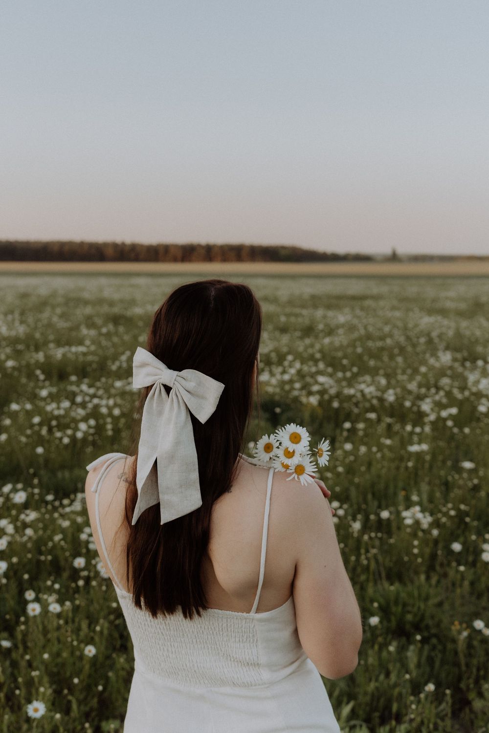 Person wearing white dress holds daisies while standing in a blooming wildflower meadow at sunset.