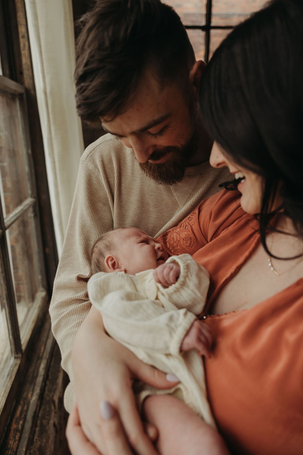 Parent in orange sweater tenderly holds and embraces swaddled infant in calm, intimate indoor setting.