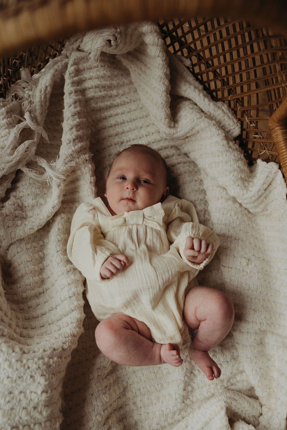Infant lying on a textured white knit blanket wearing a cream-colored dress in warm moody lighting.