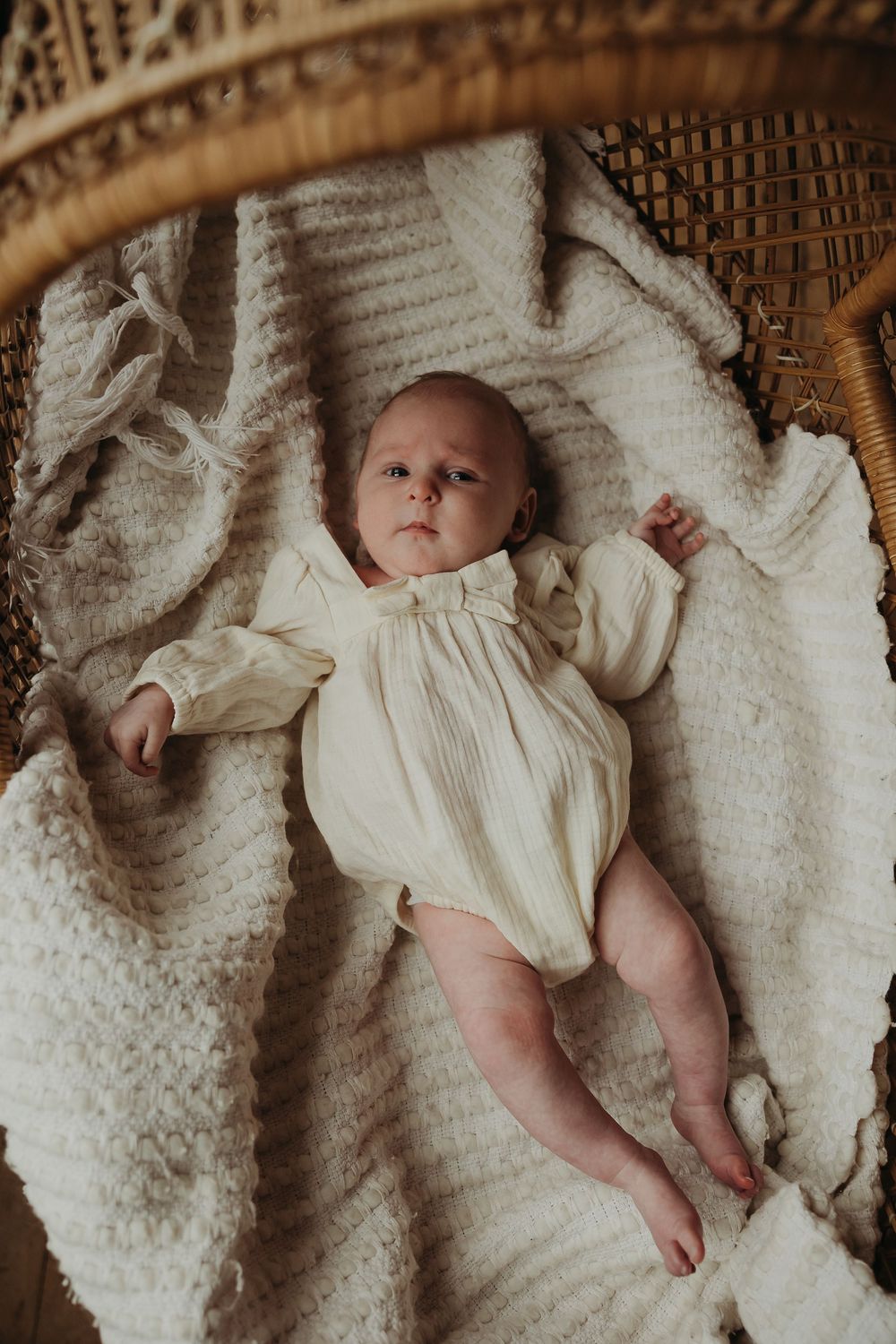 Baby wearing white vintage-style dress lying on textured cream blanket in warm sepia-toned setting.