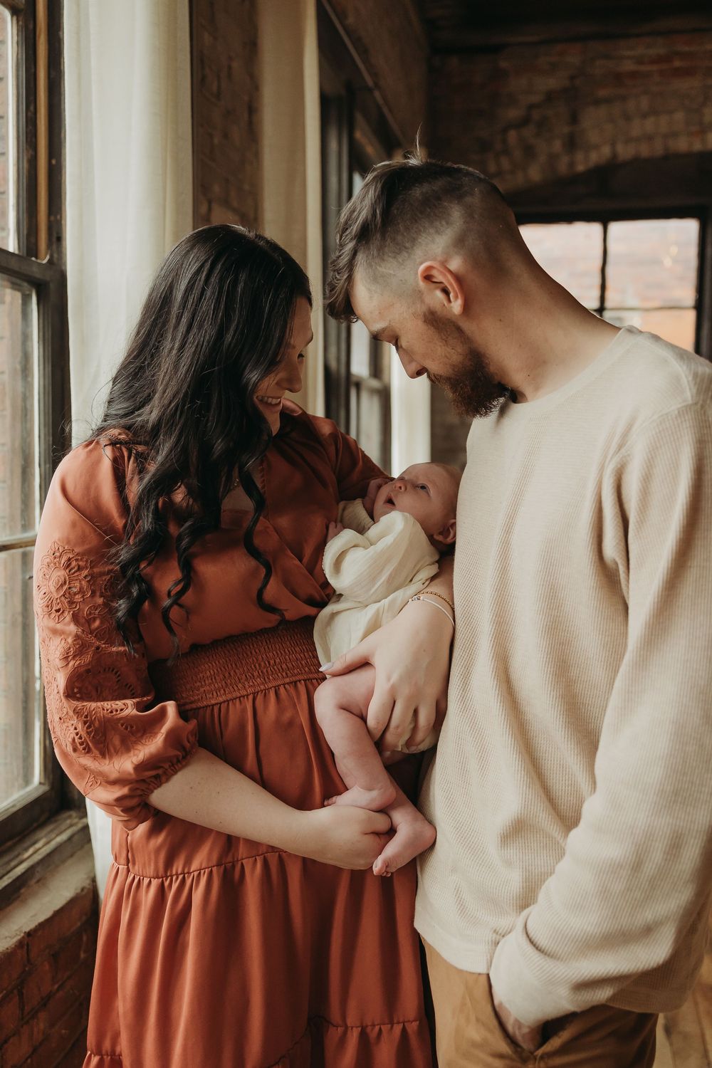 A couple in rust-colored dress and beige sweater share an intimate moment by a window while holding their newborn baby.