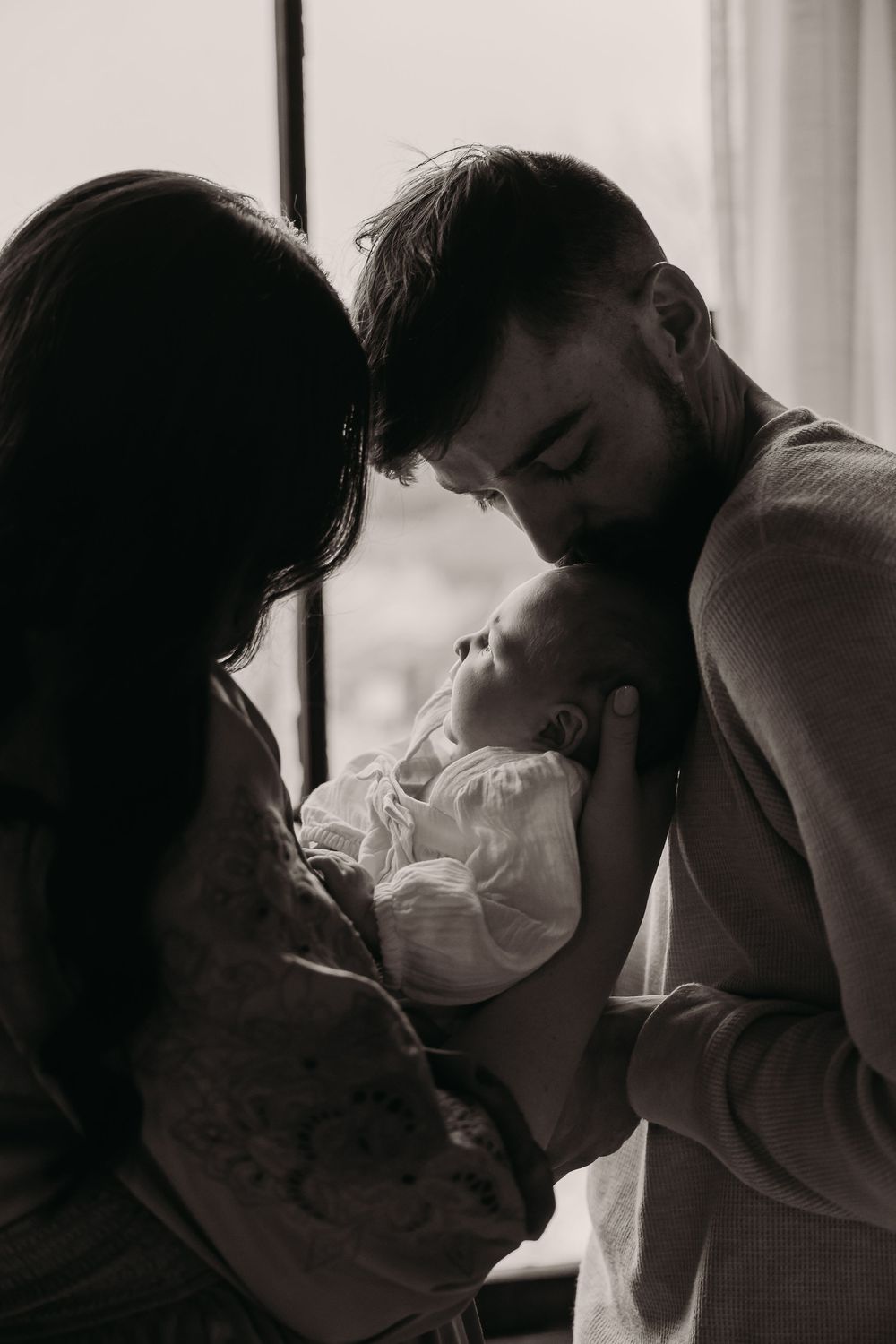 Black and white intimate family portrait showing tender moment between parents and newborn baby by window.