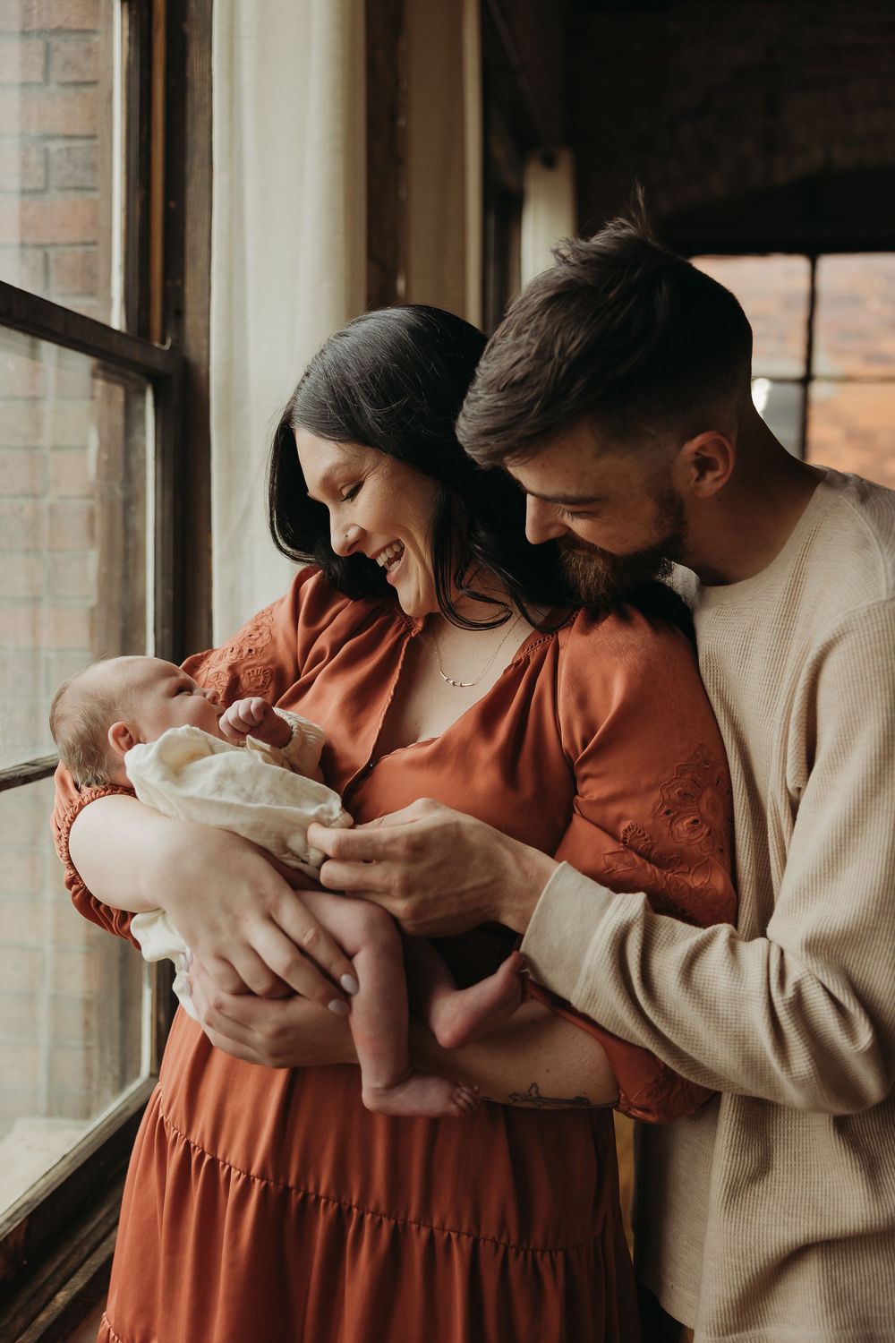 A couple wearing rust orange and beige hold and gently kiss their newborn baby in a series of intimate portraits by a window.