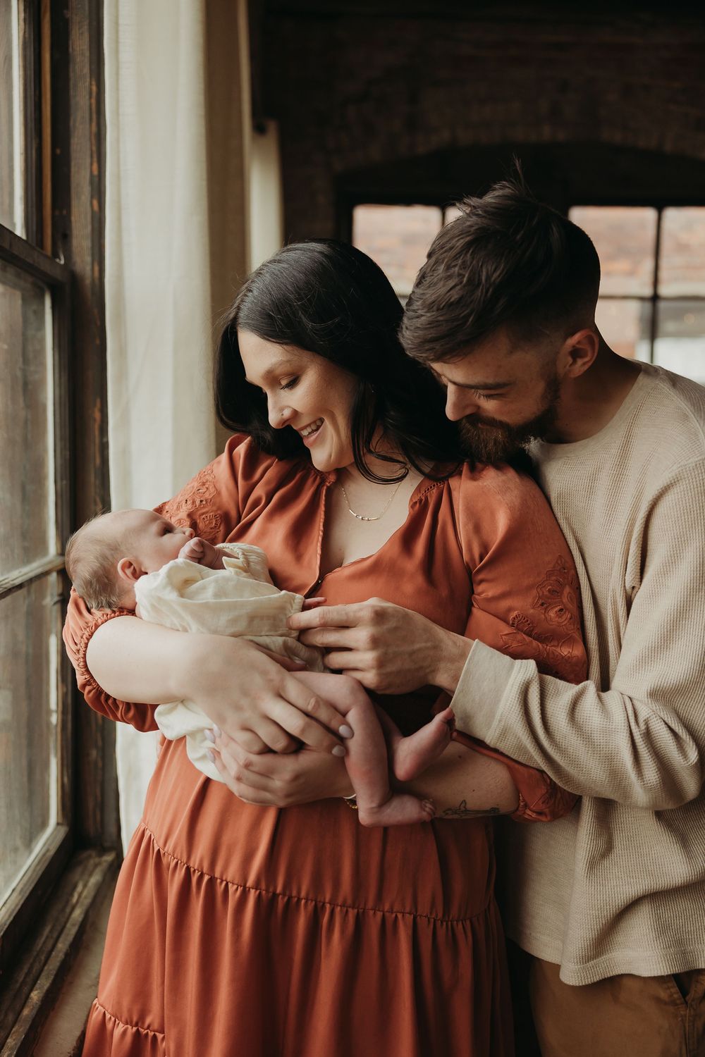 Family stands by window while lovingly gazing at their newborn baby, mother wearing rust-colored dress and father in beige sweater.